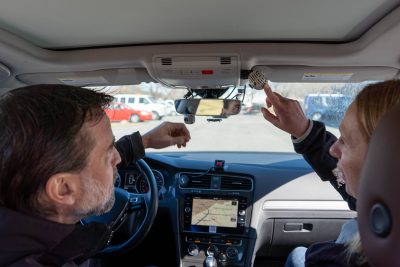 Two people sitting inside of a car pointing at a camera